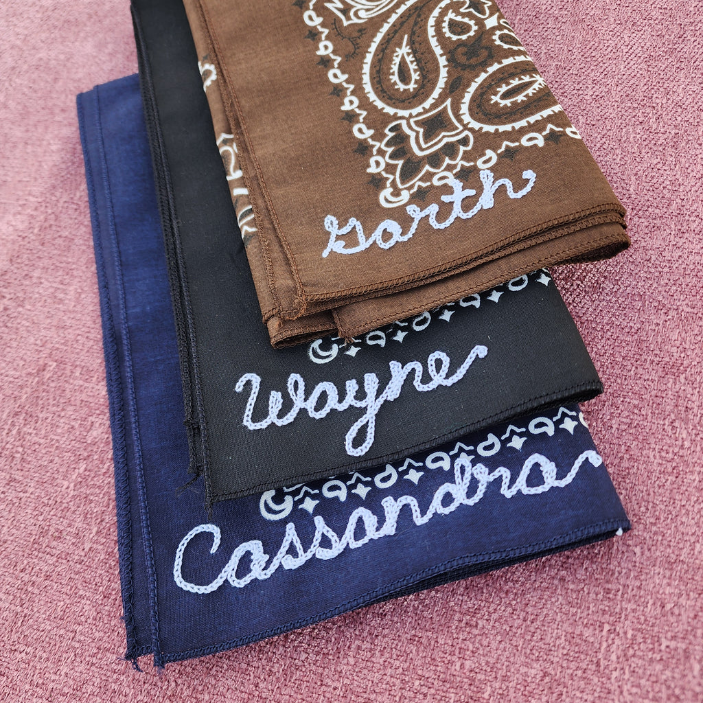 Three bandanas in brown, black, and navy blue with embroidered text that says Garth, Wayne, and Cassandra on a pink textured background
