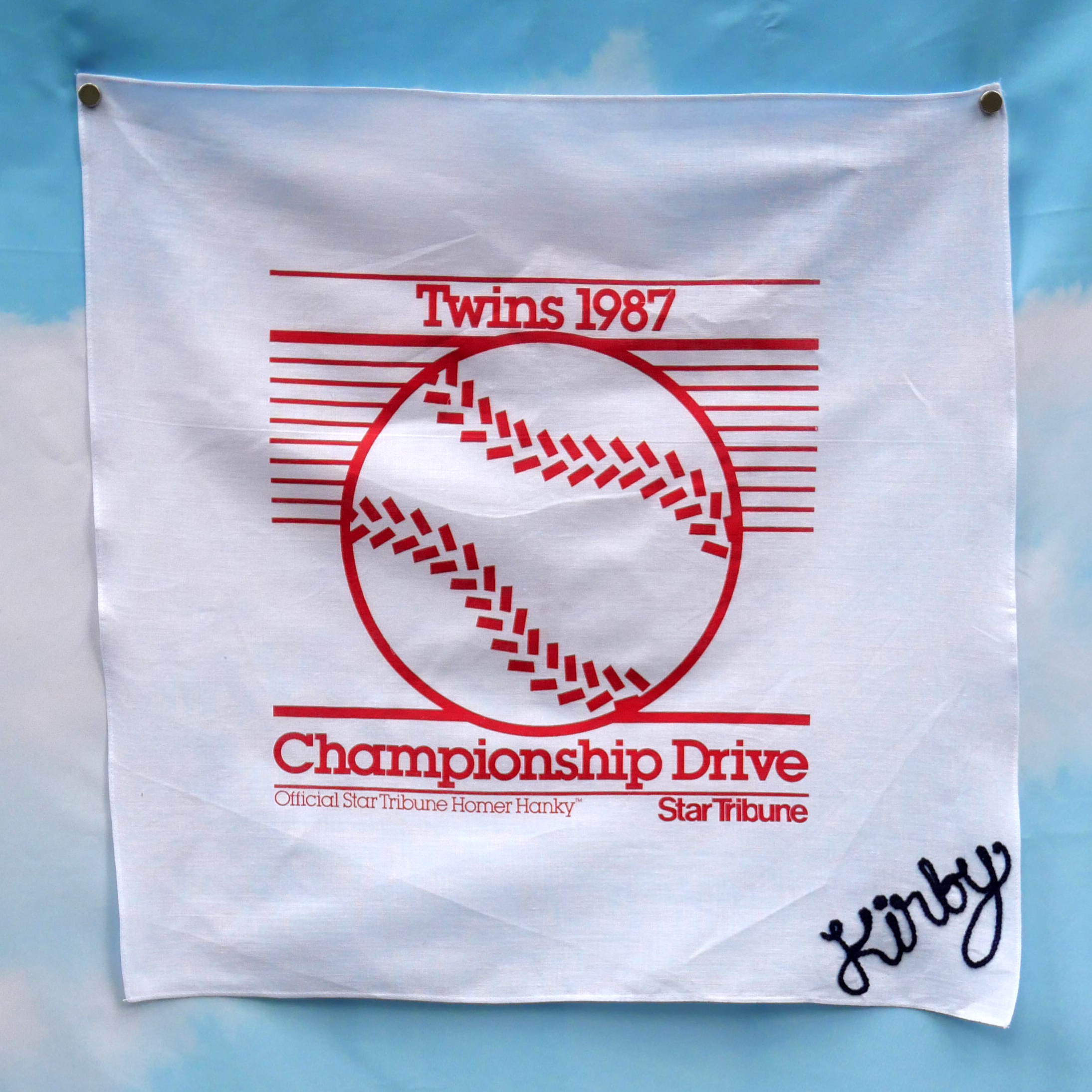 A white hankie with red text: 'Twins 1987 Championship Drive,' with 'Star Tribune' and a navy blue cursive 'Kirby' signature in the lower right corner, shown hanging against a blue sky background.